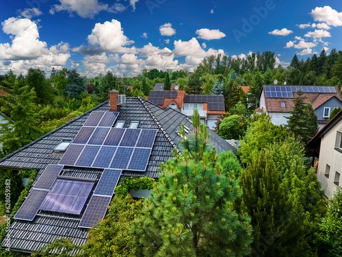 Aerial view of the roofs of houses covered with solar panels. Family houses in gardens, photovoltaic panels on the roof, summer, blue sky with white clouds. Home Electricity Generation, green living.