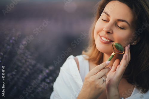 Young woman making face massage using jade roller, sitting in lavender field.