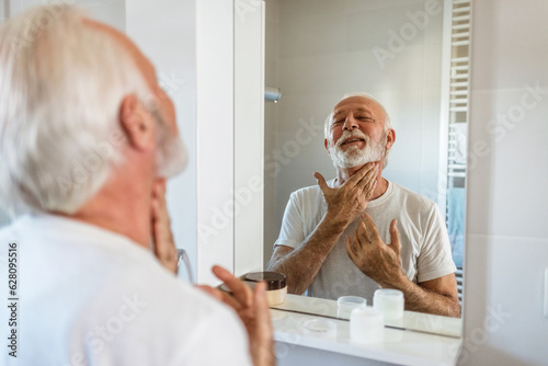 Skin care. Handsome bearded senior man applying cream at his face and looking at himself with smile while standing in front of the mirror. © Jelena Stanojkovic