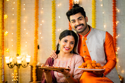 Happy smiling indian couple in traditional ethnic wear by holding sweets at home - concept of diwali festive celebration, greeting and family relationship