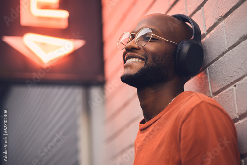 .Thinking, smile and music with a black man in the city, leaning on a brick wall on the street at night. Idea, glasses and headphones with a happy young male person streaming or listening to audio.