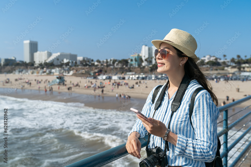 joyful stylish asian taiwanese woman tourist enjoying sunny weather and fresh air on waterfront bridge at santa monica beach with smartphone in hand