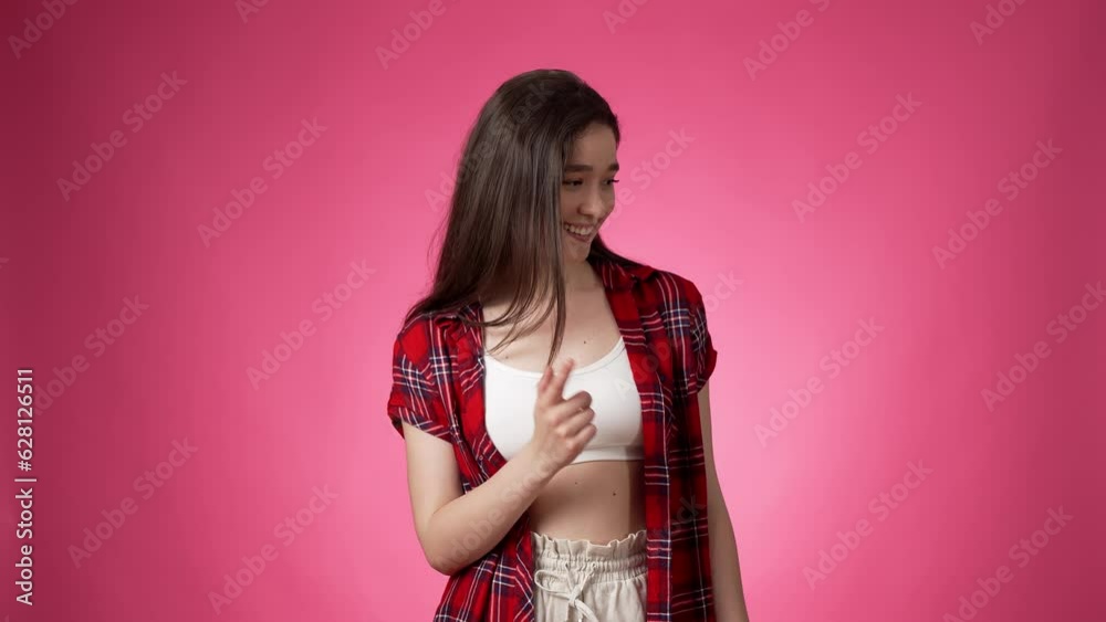 Happy young casually dressed smiling woman pointing repeatedly forefinger to side in lighthearted manner and then looking back coquettishly at camera against pink background Happy young casually dressed smiling woman pointing repeatedly forefinger to side in lighthearted manner and then looking back coquettishly at camera against pink background