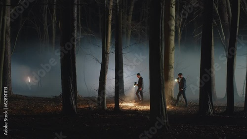 Two men walking in the night foggy forest with flashlights. Detectives searching clues or body among the trees. Crime scene, TV or movie experience, police work