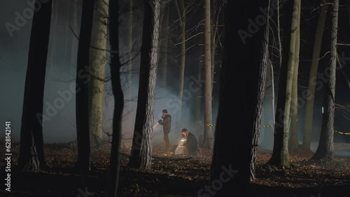 Detectives or criminologists with flashlights working on restricted crime scene in night autumn woods with dense mist. Police line, lamp lighting, flying drone. Cinematic thriller scene