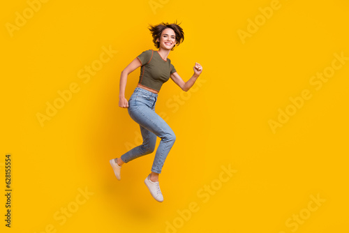 Full length photo of adorable excited girl dressed khaki top jumping high running empty space isolated yellow color background