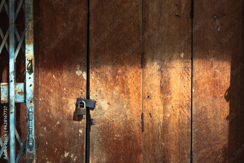 Old gold steel lock and damaged and rusted steel door on wooden door. Locked on wooden door with old steel metal shutter and folding door gate. Old wooden door with locked key. Vintage background. Old gold steel lock and damaged and rusted steel door on wooden door. Locked on wooden door with old steel metal shutter and folding door gate. Old wooden door with locked key. Vintage background.