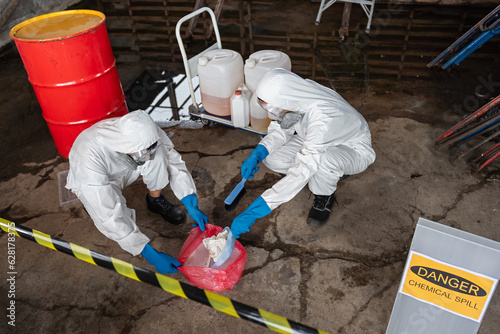 Fotografie, Obraz A team of two chemists, wearing PPE suits and gas masks, recover a deadly chemical spill on the factory warehouse floor