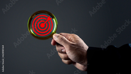 Close-up hand of businessman holding magnifier glass with dartboard and arrow for focus and setup business objective target and goal concept against dark background.