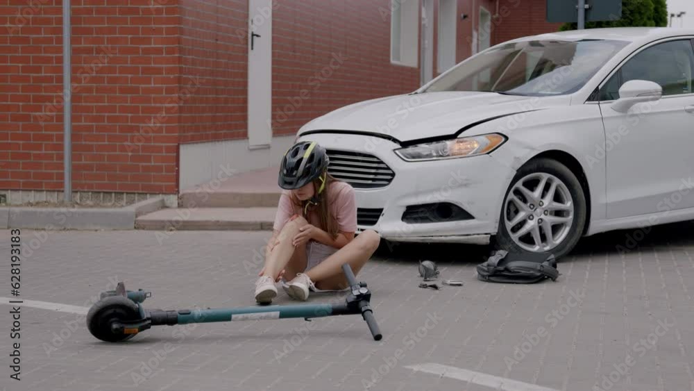 Sad young woman with protective helmet on her head, sitting on the asphalt, suffering from knee pain,rubbing her injured leg after an accident with an electric scooter and a car.protection of cyclists