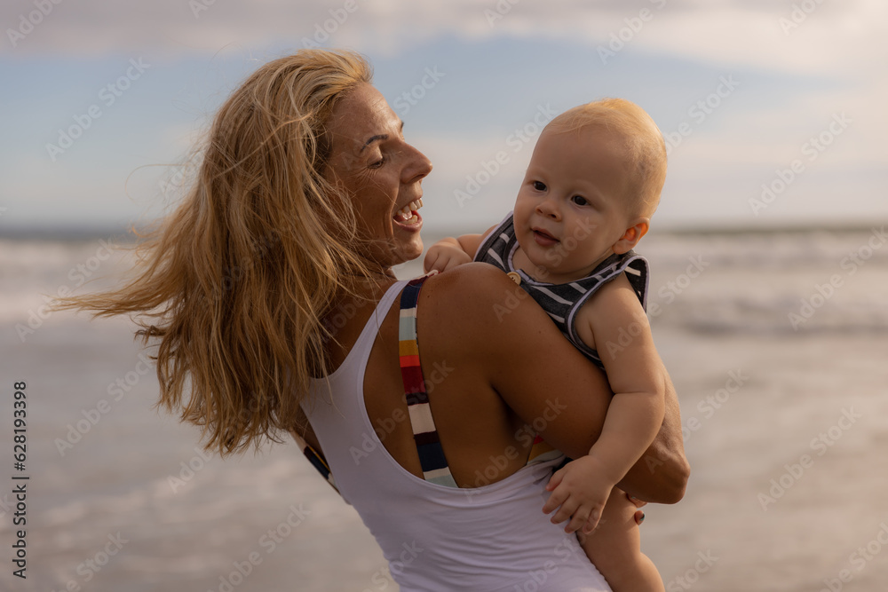 Smiling young mum and baby son. Mother with infant spending time on the beach. Summer vacation in Asia. Family concept. Happy childhood. Sunset time. Sky and ocean. Seminyak, Bali Smiling young mum and baby son. Mother with infant spending time on the beach. Summer vacation in Asia. Family concept. Happy childhood. Sunset time. Sky and ocean. Seminyak, Bali