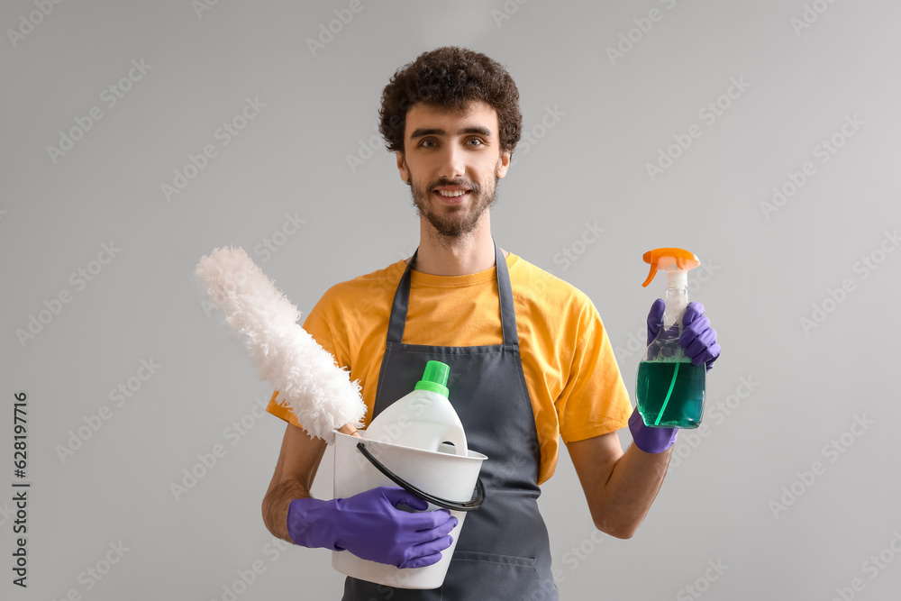 Young man with bucket of cleaning supplies on grey background Young man with bucket of cleaning supplies on grey background