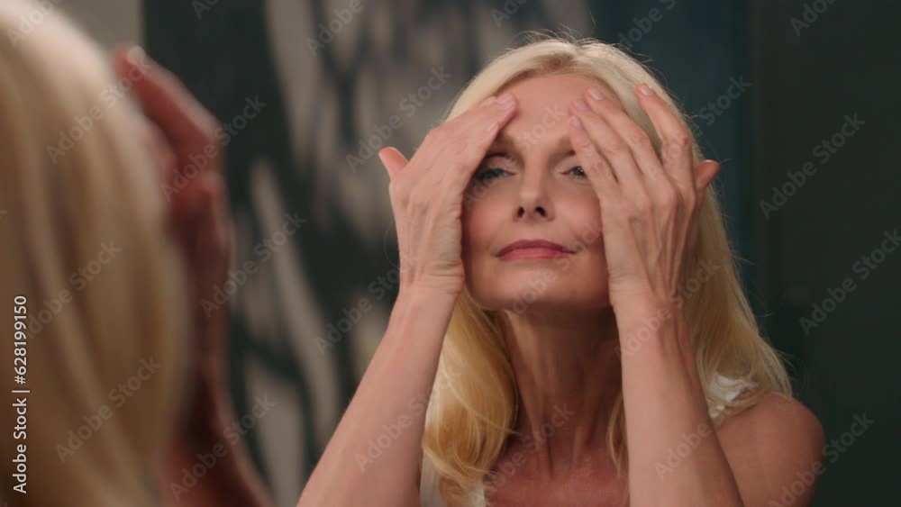 Mature senior Caucasian 50s woman touching perfect face looking at mirror applying sunscreen anti wrinkle age lifting moisturizing cream with collagen skincare tighten skin rejuvenation beauty routine