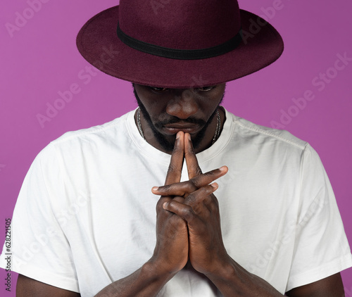Thoughtful young black man standing in hat near purple wall with fingers near lips