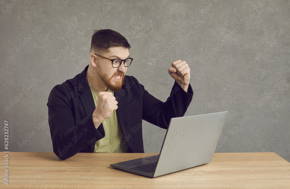 Funny man having problem with computer. Angry furious crazy young guy in glasses sitting at desk, clenching fists and looking at laptop screen threatening to hit and break it. Software error concept Funny man having problem with computer. Angry furious crazy young guy in glasses sitting at desk, clenching fists and looking at laptop screen threatening to hit and break it. Software error concept