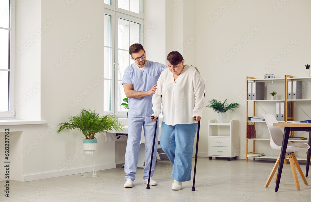 Male caregiver helping his female patient with a leg injury. Young man in a uniform supports an overweight woman while she walks with crutches in a medical room at the clinic or hospital Male caregiver helping his female patient with a leg injury. Young man in a uniform supports an overweight woman while she walks with crutches in a medical room at the clinic or hospital