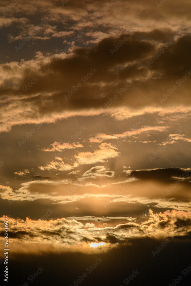 large orange clouds in the blue sky, during the golden hour, through which the bright rays of the sun break through large orange clouds in the blue sky, during the golden hour, through which the bright rays of the sun break through