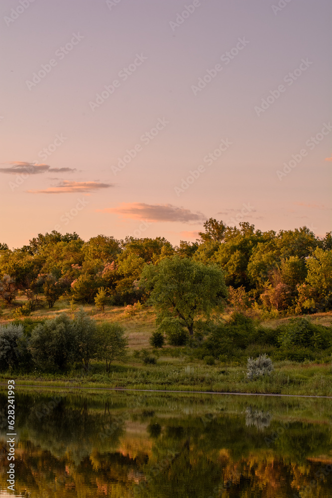 fields with hills on which green grass, bushes and trees grow, near a calm lake, against the backdrop of a bright sunset sky with clouds, during the golden hour fields with hills on which green grass, bushes and trees grow, near a calm lake, against the backdrop of a bright sunset sky with clouds, during the golden hour