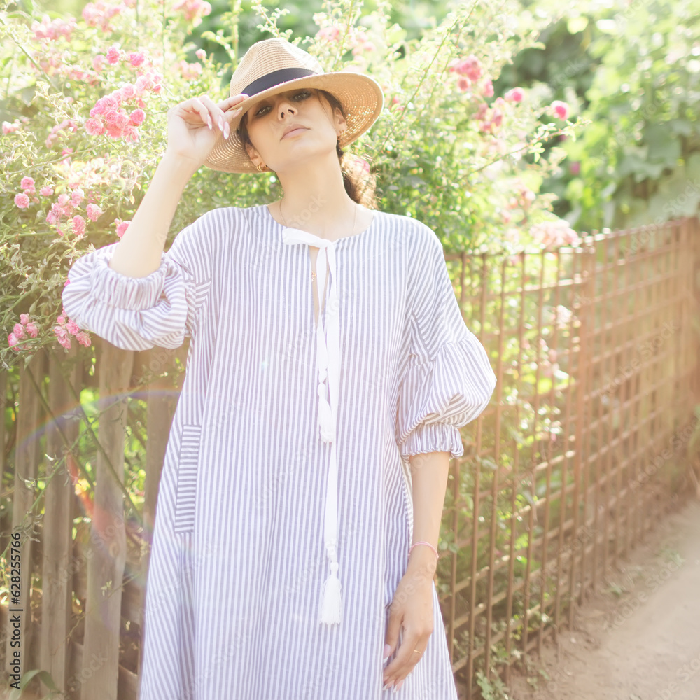 Romantic stylishly dressed young adult beautiful woman relaxing in a countryside park looking away. Blonde Parisian-styled model in a white transparent dress and straw hat walking against of garden