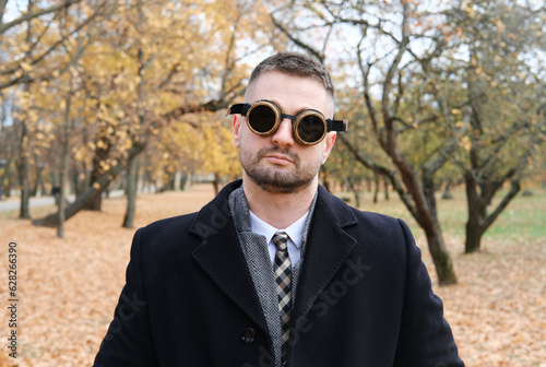 A man in a business suit, coat and steampunk goggles poses in an autumn park