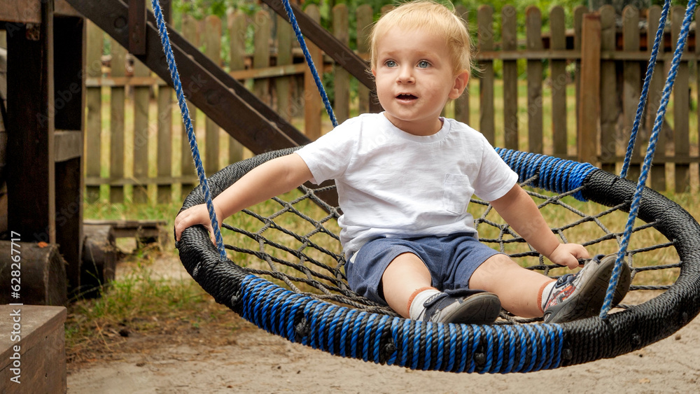 Little baby boy having fun and smiling while riding in rope nest swing at park. Kids playing outdoors, children having fun, summer vacation Little baby boy having fun and smiling while riding in rope nest swing at park. Kids playing outdoors, children having fun, summer vacation