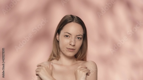 The woman crosses her arms and touches the bare skin of her shoulders. Portrait of an attractive seminude woman in the studio on a pink background close up. The concept of beauty, cosmetology and care