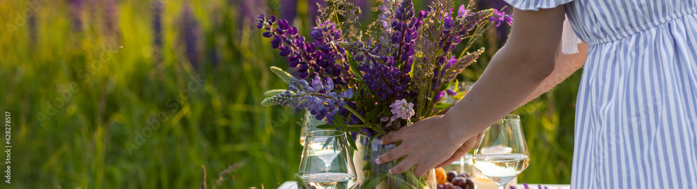 Beautiful happy young woman on a meadow arranging table for outdoor event, gathering wildflowers, lighting candles. Wedding or romantic date decoration in the field with purple lupins, fruits banner