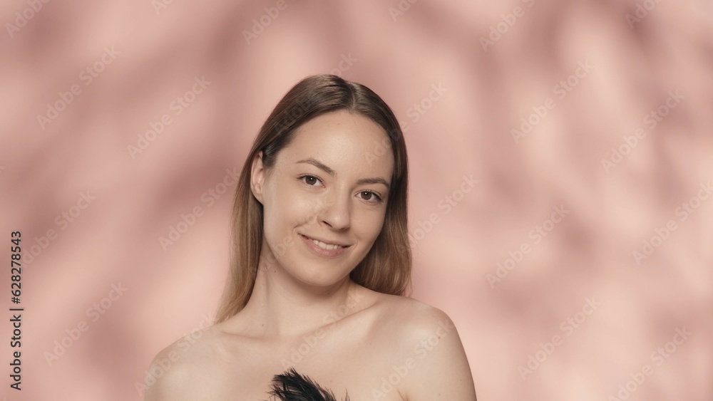 Portrait of a smiling seminude woman on a pink background close up. A woman with natural makeup looks straight ahead, holding a black feather. Natural beauty, cosmetology, skin care. Portrait of a smiling seminude woman on a pink background close up. A woman with natural makeup looks straight ahead, holding a black feather. Natural beauty, cosmetology, skin care.