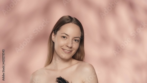 Portrait of a smiling seminude woman on a pink background close up. A woman with natural makeup looks straight ahead  holding a black feather. Natural beauty  cosmetology  skin care.