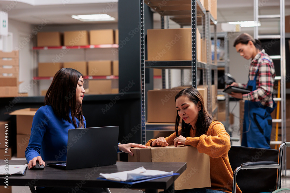 Warehouse delivery manager checking parcel packing quality while working together with order picker in wheelchair. Storehouse shipment operators preparing cardboard box with goods for distribution