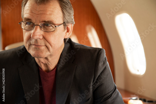 Portrait of mature business man wearing glasses sitting in seat aboard private jet looking off camera 