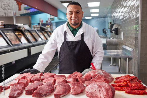 Smiling portrait of butcher wearing hair net behind counter of meat market of grocery store with selection of cuts of steak  photo