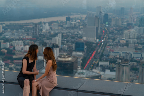 Asian woman friends meeting and looking urban cityscape together at skyscraper rooftop cafe bar in metropolis at summer sunset. Attractive girl relax and enjoy urban lifestyle in the city at night.