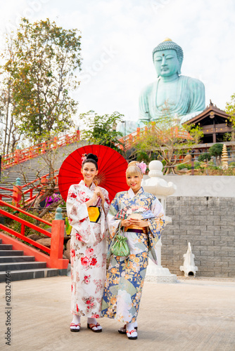 Vertical image of two Asian women wear japanese style dress with one hold red umbrella and stand in front of stair to green big buddha statue. © narong