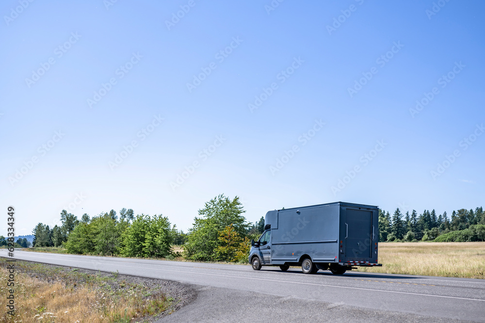 Small rig semi truck with huge box trailer making delivery running on the narrow winding road with summer meadows on the sides