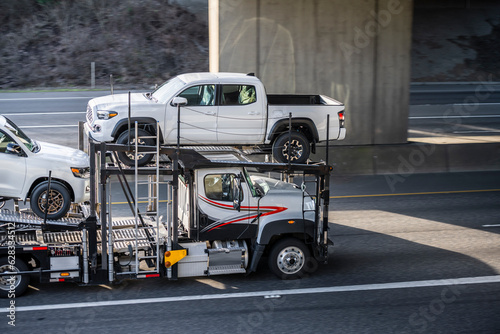 White industrial standard big rig car hauler semi truck transporting pick up trucks on two level semi trailer driving on the highway road under the bridge