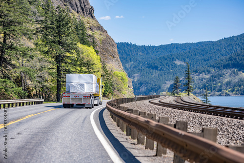  Yellow classic bonnet big rig semi truck transporting covered cargo on flat bed semi trailer driving on the winding highway road along the Columbia River and parallel to the railroad