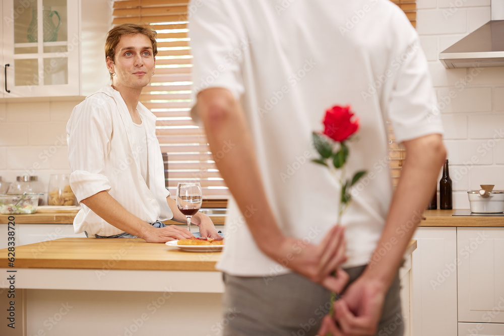 young gay couple looking at boyfriend and holding rose behind his back for surprise in the kitchen young gay couple looking at boyfriend and holding rose behind his back for surprise in the kitchen