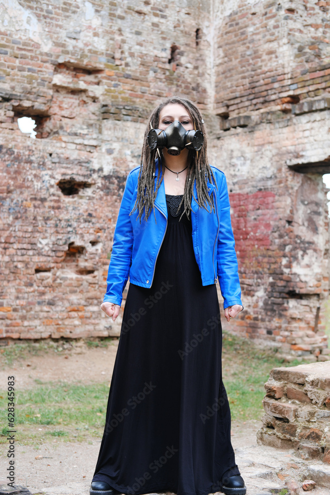 Naklejka premium Woman wearing gas mask with spikes in blue jacket and black dress standing in ruined building
