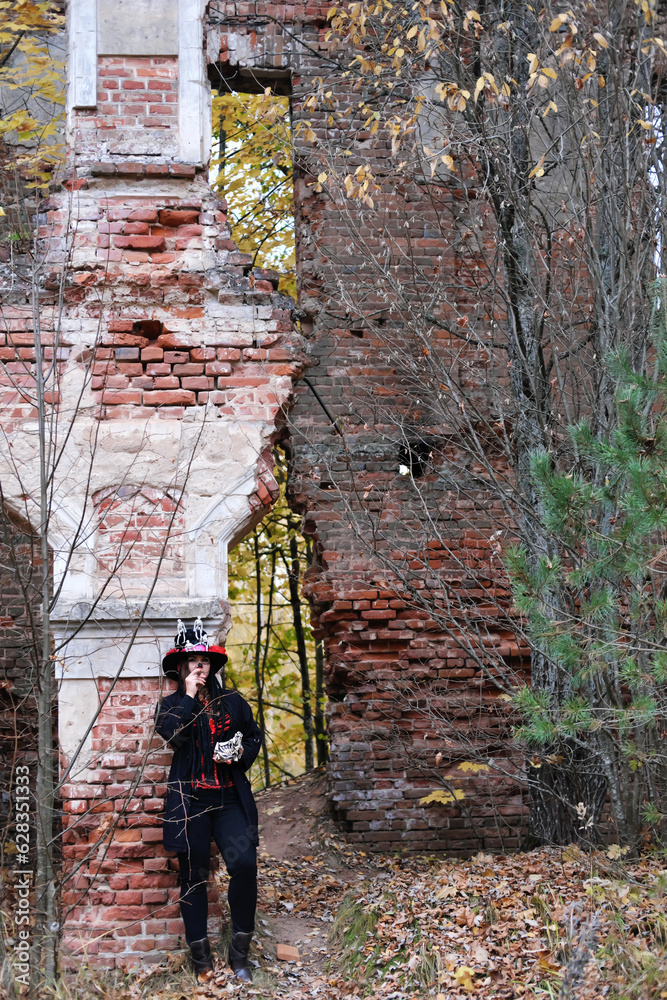 A woman in the image of Baron Saturday poses against the backdrop of an old abandoned building with a cigar in her hands. The model is dressed in a corset, a cardigan and a top hat 

