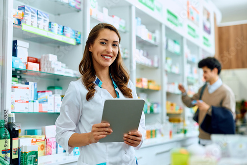 Happy female pharmacist with touchpad working in pharmacy and looking at camera.