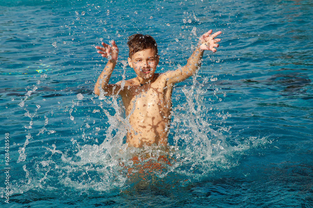 The boy is playing in the pool. The child raises his hands and splashes water. Games in the outdoor pool. Splashing and swimming in the sea, healthy lifestyle, summer holidays.
