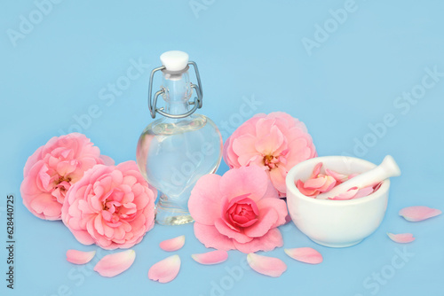 Rose flower perfume in heart shaped bottle with flowers and loose petals on blue. Preparation of scented floral product, gift for Valentines Day, birthday, anniversary or Mothers day.