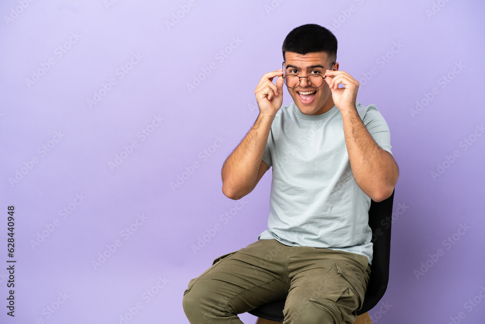 Young man sitting on a chair over isolated background with glasses and surprised Young man sitting on a chair over isolated background with glasses and surprised