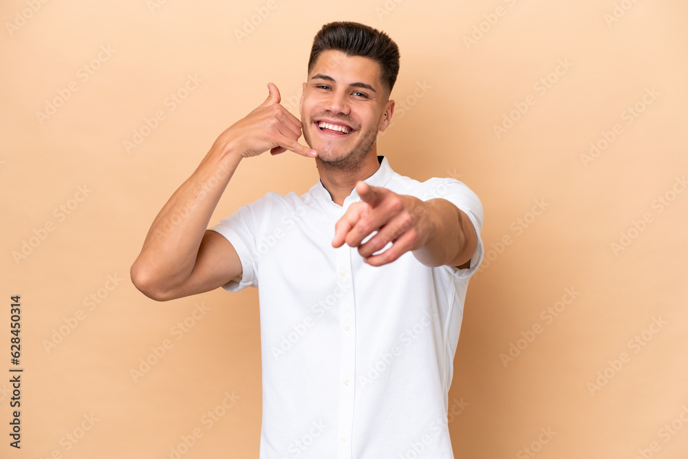 Young caucasian man isolated on beige background making phone gesture and pointing front