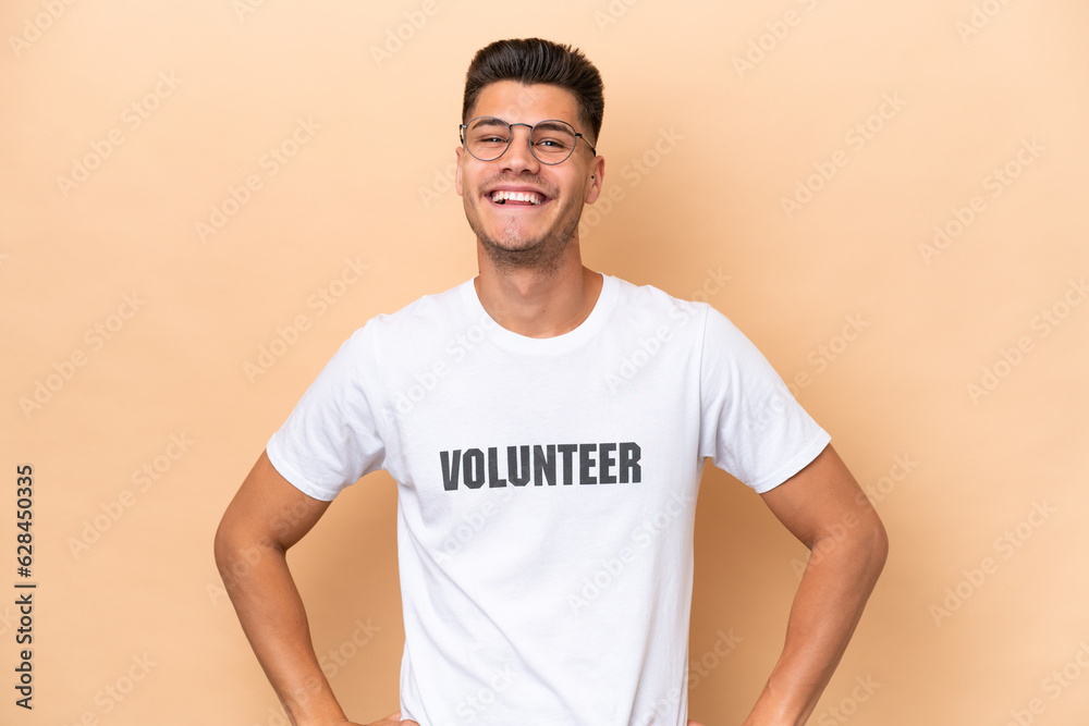 Young volunteer caucasian man isolated on beige background posing with arms at hip and smiling Young volunteer caucasian man isolated on beige background posing with arms at hip and smiling