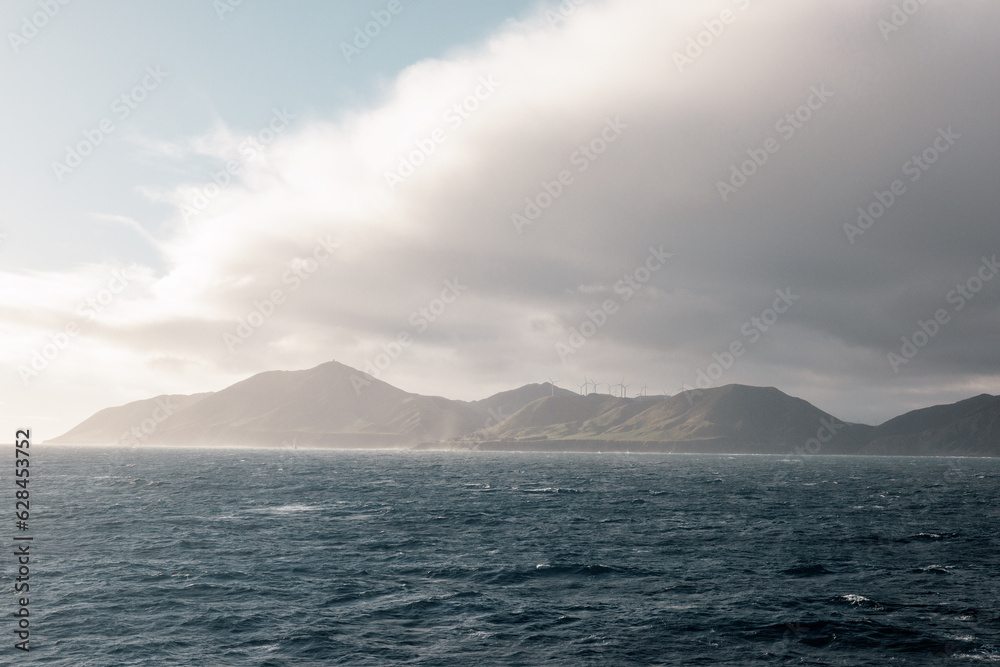 A rugged and moody looking coastline.  The coast is in Wellington New Zealand. On the hills are a number of windmills