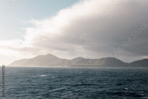 A rugged and moody looking coastline. The coast is in Wellington New Zealand. On the hills are a number of windmills