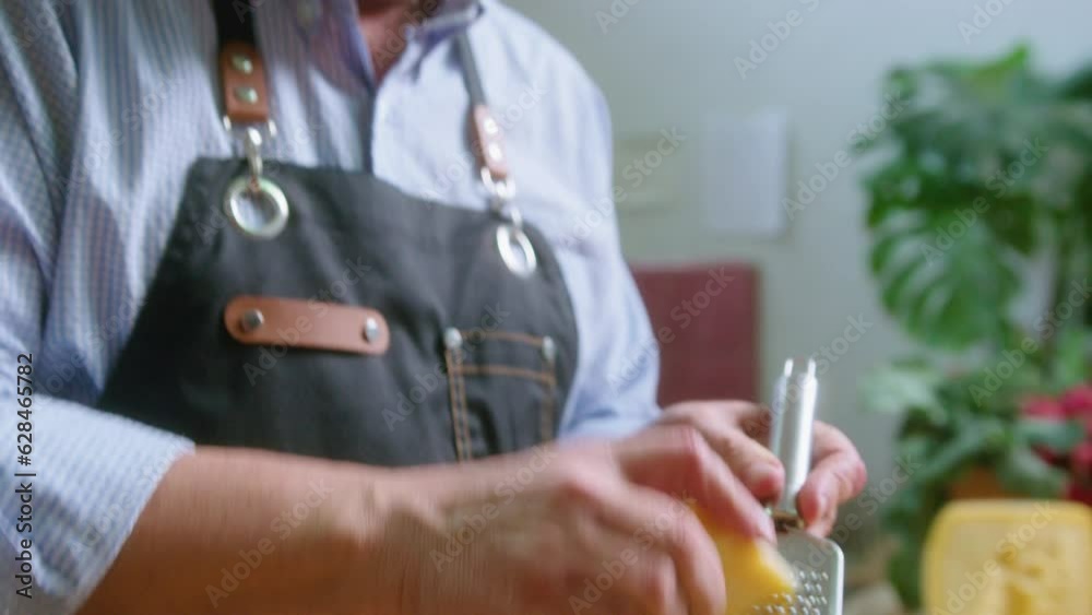 Senior food blogger in apron adding grated cheese to salad in bowl, looking at camera and speaking while recording video recipe in the kitchen. Handheld shot