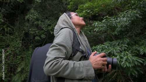 Solo Asian woman nature serious photographer holding camera device looking for stunning shot in the lush foliage rainforest. Outdoor activities. Beauty in nature. Travel destination.